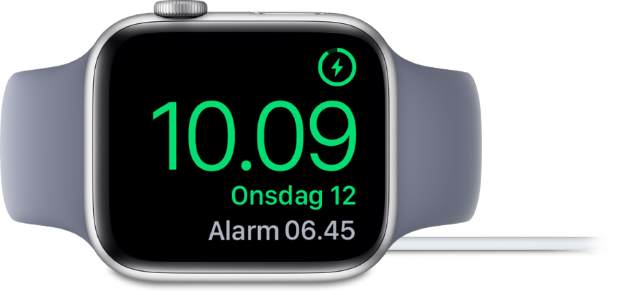 Et Apple Watch, der ligger på siden og er sluttet til opladeren, hvor skærmen viser opladersymbolet øverst til højre, det aktuelle klokkeslæt derunder og klokkeslættet for den næste alarm.