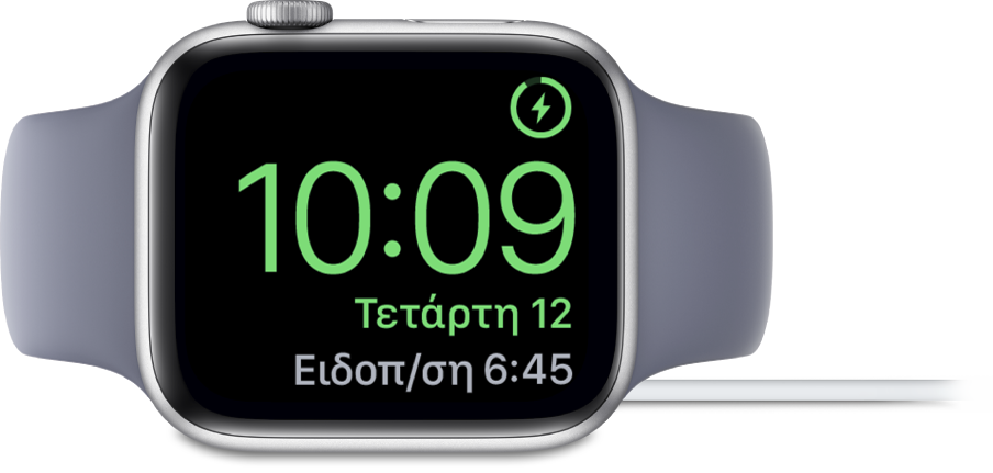 Ένα Apple Watch τοποθετημένο στο πλάι του και συνδεδεμένο στον φορτιστή του. Στην οθόνη εμφανίζεται το σύμβολο φόρτισης στην πάνω δεξιά γωνία, η τρέχουσα ώρα από κάτω η ώρα του επόμενου ξυπνητηριού.