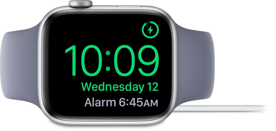 An Apple Watch placed on its side and connected to the charger, with the screen showing the charging symbol in the top-right corner, the current time below that, and the time of the next alarm.