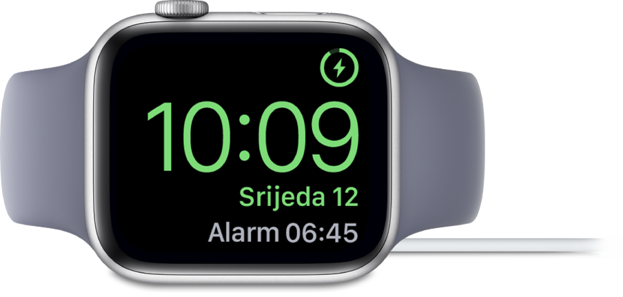 Apple Watch postavljen na bok i spojen na punjač, na čijem zaslonu je u gornjem desnom kutu prikazan simbol napajanja, trenutačno vrijeme dolje ispod i vrijeme sljedećeg alarma.