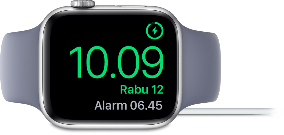 Apple Watch diletakkan di sampingnya dan tersambung ke pengisi daya, dengan layar menampilkan simbol pengisian daya di pojok kanan atas, waktu saat ini di bawahnya, dan waktu alarm berikutnya.
