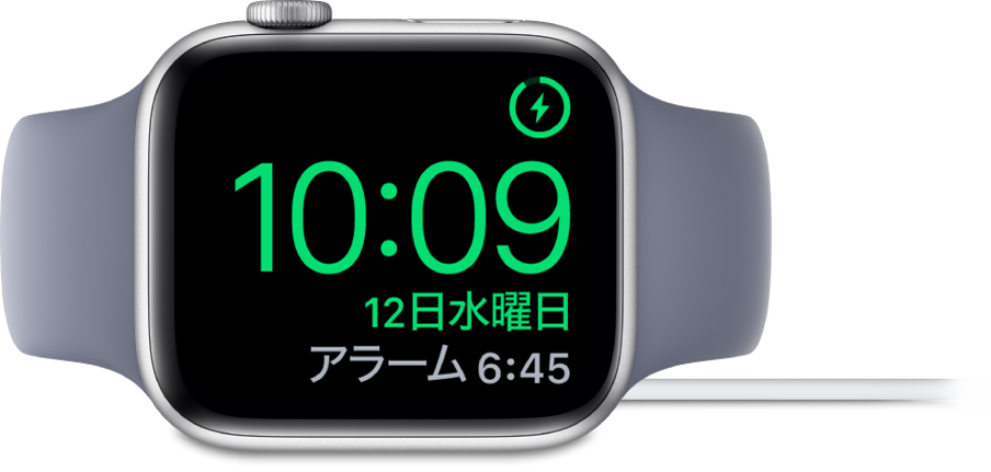 横向きに置かれ、充電器に接続されているApple Watch。画面には、右上隅に充電中のマーク、その下に現在時刻と、次のアラーム時刻が表示されています。