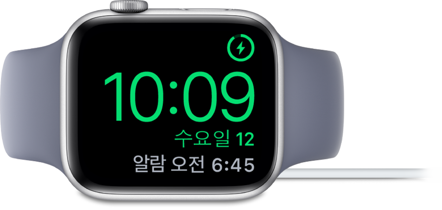 Apple Watch가 옆으로 누워있고 충전기에 연결되어 있습니다. 화면의 오른쪽 상단 모서리에는 충전 기호가 있고 그 아래에는 현재 시간과 다음 알람 시간이 있습니다.