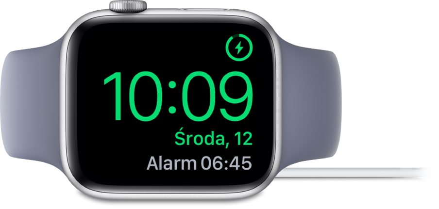 Apple Watch ustawiony na boku i podłączony do ładowarki. W prawym górnym rogu ekranu widoczny jest symbol ładowania, niżej wyświetlana jest bieżąca godzina, a dalej znajduje się czas najbliższego alarmu.