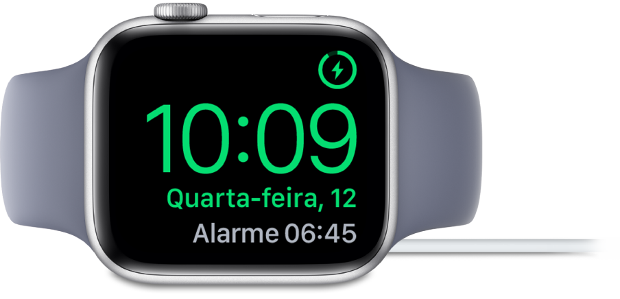 Apple Watch posicionado de lado e conectado ao carregador, com a tela mostrando o símbolo de carregamento no canto superior direito, a hora atual abaixo e o horário do próximo alarme.