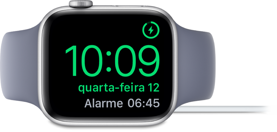 Um Apple Watch colocado de lado e ligado ao carregador, com o ecrã a mostrar o símbolo de carregamento no canto superior direito e, por baixo, a hora atual e a hora do próximo alarme.