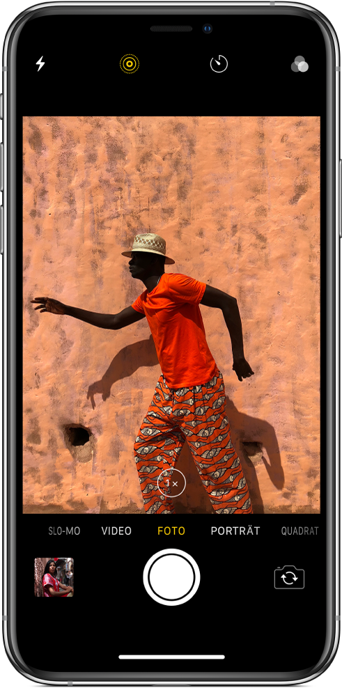 Kamera im Modus „Foto“ mit den anderen Modi links und rechts unter der Bildansicht. Oben auf dem Bildschirm sind die Tasten „Blitz“, „Live Photo“, „Timer“ und „Filter“ zu sehen. Durch Tippen auf das Miniaturbild unten links kannst du auf vorhandene Fotos und Videos zugreifen. In der Mitte unten befindet sich die Taste „Auslöser“ und rechts unten die Taste „Kamera wechseln“.