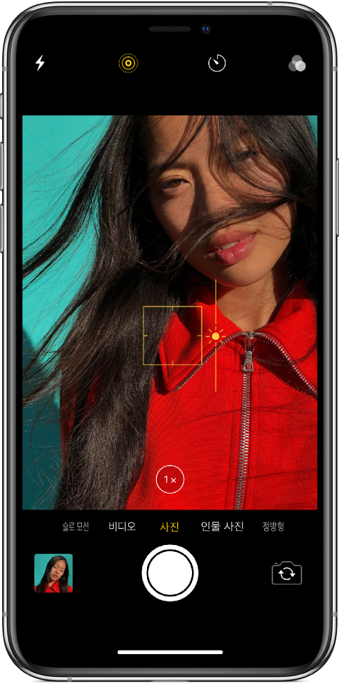 사진 모드 상태의 카메라 앱 화면. 뷰어에서 노란색 상자 윤곽선은 초점 영역을 표시하며 슬라이더를 위 또는 아래로 드래그하여 노출을 조절할 수 있습니다. 확대에 1x 확대/축소 버튼을 사용할 수 있습니다.