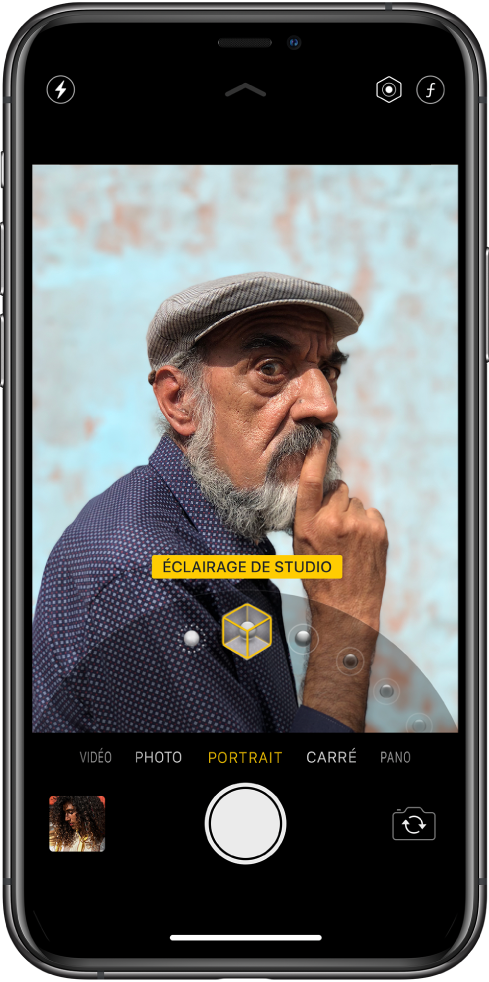 L’écran « Appareil photo » en mode Portrait ; le sujet est net tandis que l’arrière-plan est flou. Le cadran pour sélectionner les effets d’éclairage Portrait est ouvert en bas de l’image et « Éclairage de studio » est sélectionné. En haut à gauche de l’écran se trouve le bouton Flash, et en haut à droite de l’écran se trouvent les boutons permettant de régler l’intensité lumineuse du portrait et le contrôle de la profondeur. Près du bas de l’écran figurent, de gauche à droite, une vignette permettant d’accéder aux photos et vidéos, le bouton Obturateur et le bouton « Changer de caméra ».