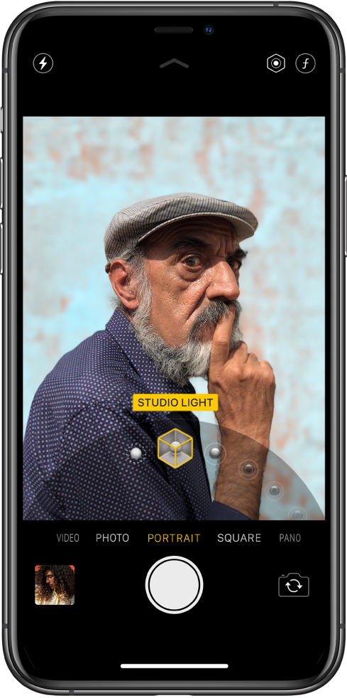 Portreto režimu veikiančios programos „Camera“ ekranas; objektas ryškus, o fonas sulietas. Kadro apačioje atidarytas portreto apšvietimų efekto parinkiklis ir nustatyta studijinio apšvietimo parinktis. Ekrano viršutiniame kairiajame kampe yra blykstės mygtukas, viršutiniame dešiniajame kampe – portreto apšvietimo intensyvumo koregavimo mygtukas ir gylio valdiklis. Kameros režimų apačioje (iš kairės į dešinę) yra vaizdo miniatiūra, kad pasiektumėte nuotraukas ir vaido įrašus, „Shutter“ mygtukas ir „Switch Camera“ mygtukas.
