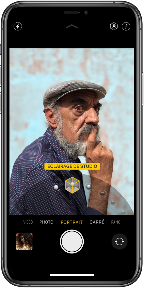 L’écran « Appareil photo » en mode Portrait ; le sujet est net tandis que l’arrière-plan est flou. Le cadran pour sélectionner les effets d’éclairage Portrait est ouvert en bas de l’image et « Éclairage de studio » est sélectionné. En haut à gauche de l’écran se trouve le bouton Flash, et en haut à droite de l’écran se trouvent les boutons permettant de régler l’intensité lumineuse du portrait et le contrôle de la profondeur. Près du bas de l’écran figurent, de gauche à droite, une vignette permettant d’accéder aux photos et vidéos, le bouton Obturateur et le bouton « Changer de caméra ».
