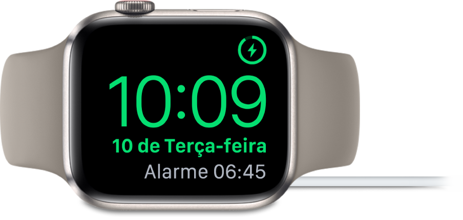 Apple Watch posicionado de lado e conectado ao carregador, com a tela mostrando o símbolo de carregamento no canto superior direito, a hora atual abaixo e o horário do próximo alarme.