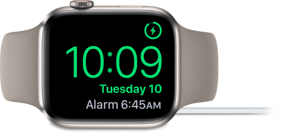 Apple Watch, поставен на една страна и свързан към зарядното устройство, екранът показва символа за зареждане в горния десен ъгъл, текущия час под него и часа на следващата аларма.