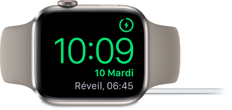 Apple Watch placée sur sa tranche et connectée au chargeur. L’écran montre le symbole de recharge dans le coin supérieur droit, l’heure actuelle en dessous et l’heure du prochain réveil.