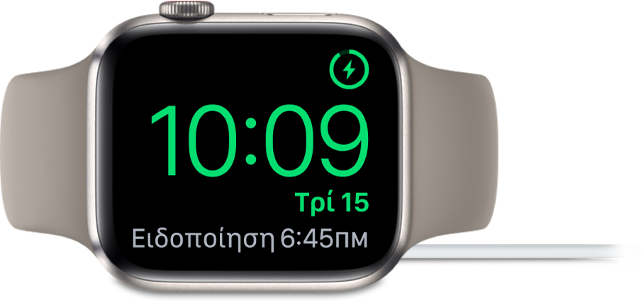 Ένα Apple Watch τοποθετημένο στο πλάι του και συνδεδεμένο στον φορτιστή του. Στην οθόνη εμφανίζεται το σύμβολο φόρτισης στην πάνω δεξιά γωνία, η τρέχουσα ώρα από κάτω η ώρα του επόμενου ξυπνητηριού.