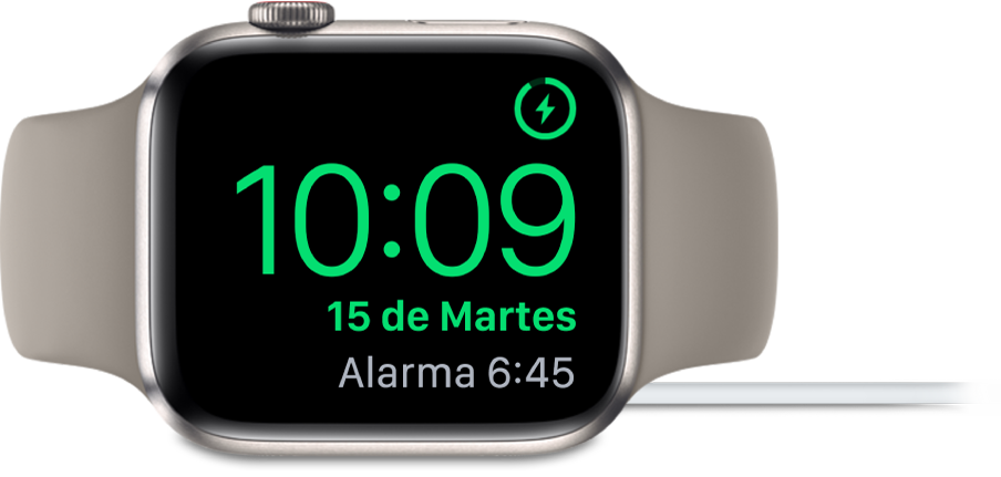 Un Apple Watch colocado de lado y conectado al cargador, en cuya pantalla se muestra el símbolo de carga en la esquina superior derecha, la hora actual debajo y la hora de la siguiente alarma.