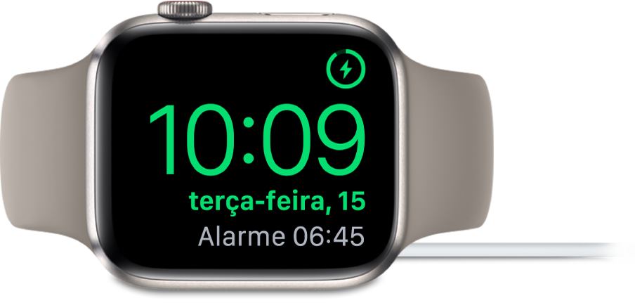 Um Apple Watch colocado de lado e ligado ao carregador, com o ecrã a mostrar o símbolo de carregamento no canto superior direito e, por baixo, a hora atual e a hora do próximo alarme.
