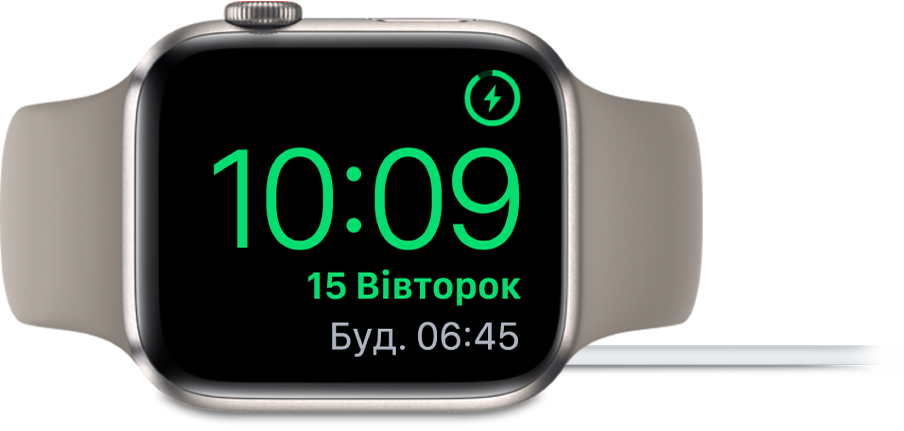Apple Watch кладеться на бік і підключається до зарядного пристрою. На екрані відображається значок заряджання у верхньому правому куті, під ним — поточний час і час наступного будильника.