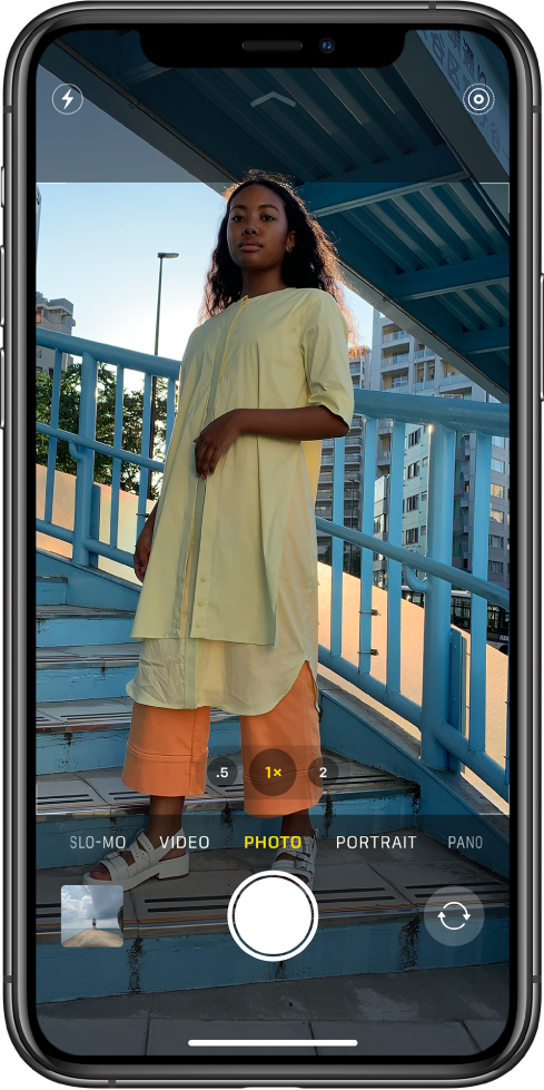 Camera in Photo mode, with other modes to the left and right below the frame. Buttons for Flash, Night Mode, and Live Photo are at the top of the screen. Above the camera modes are buttons to zoom in and out. Below the camera modes are, from left to right, an image thumbnail to access photos and videos, the Shutter button, and the Switch Camera button.