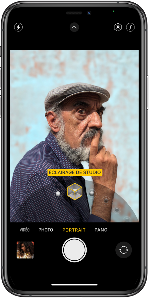 L’écran « Appareil photo » en mode Portrait ; dans le viseur, le sujet est net tandis que l’arrière-plan est flou. Le cadran pour sélectionner les effets d’éclairage Portrait est ouvert en bas de l’image et « Éclairage de studio » est sélectionné. En haut à gauche de l’écran se trouve le bouton Flash, en haut au centre se trouve le bouton « Commandes de l’appareil photo », et en haut à droite de l’écran se trouvent les boutons permettant de régler l’intensité lumineuse du portrait et le contrôle de la profondeur. En bas de l’écran, de gauche à droite, se trouvent le bouton « Visualiseur de photos et vidéos », le bouton « Prendre une photo » et le bouton « Sélecteur de caméra - face arrière ».