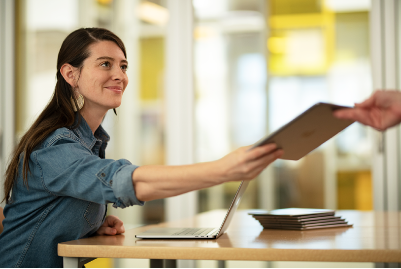 Une femme assise à son bureau dans une classe, avec un ordinateur portable Mac ouvert et une pile d’iPad à proximité. Elle remet un iPad à un élève.