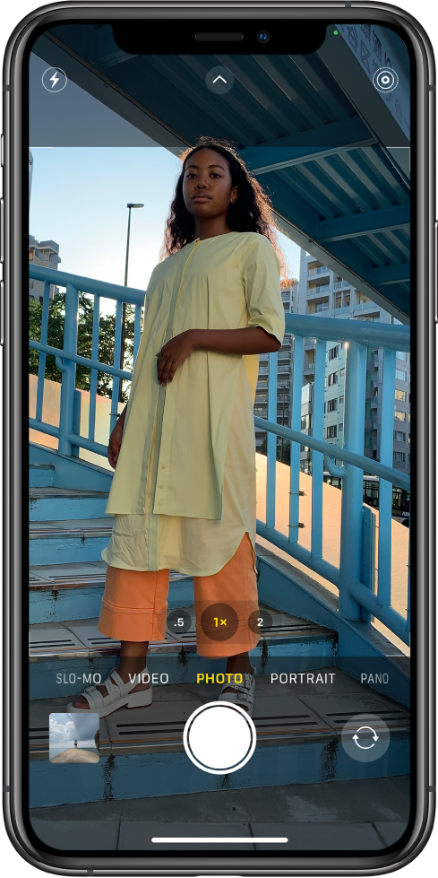 Camera in Photo mode, with other modes to the left and right below the view finder. Buttons for Flash, Night mode, Camera Controls, and Live Photo appear at the top of the screen. The Photo and Video Viewer button is in the bottom-left corner. The Take Picture button is at the bottom center, and the Camera Chooser Back-Facing button is in the bottom-right corner.