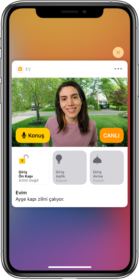 iPhone ekranında Ev uygulamasından bir bildirim. Sol tarafta bir Konuş düğmesiyle ön kapıdaki bir kişinin resmi gösteriliyor. Alt tarafta ön kapı ve giriş ışıkları için aksesuar düğmeleri var. Aksesuar düğmelerinin alt tarafında “Ayşe kapı zilini çalıyor” sözcükleri görünüyor. Bildirimin sağ üst tarafında Kapat düğmesi var.