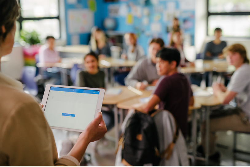 Una docente in piedi di fronte a una classe di studenti seduti, con iPad aperto su un'app educativa.