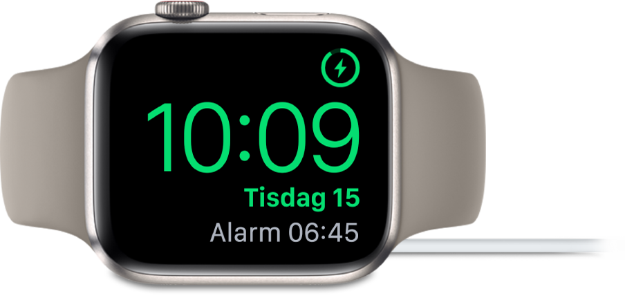 En Apple Watch som ligger på sidan och är ansluten till laddaren. Skärmen visar laddningssymbolen i det övre högra hörnet, under det tiden och sedan tiden för nästa alarm.