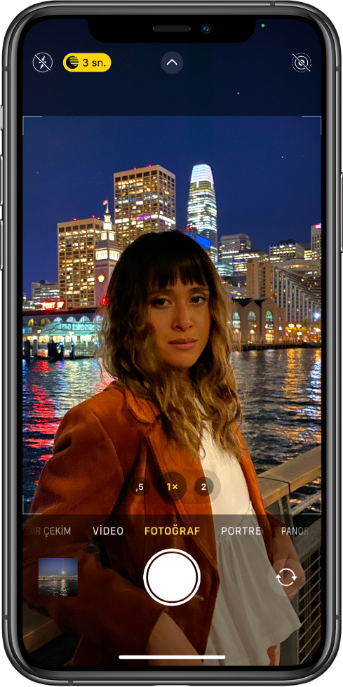 Fotoğraf modunda Kamera ekranı. Flaş ve Gece modu düğmeleri ekranın sol üstünde görünüyor. Flaş kapalı ve Gece modu açık. Kamera Denetimleri düğmesi üst ortada ve Live Photo düğmesi sağ üst köşede. Ekranın en altında soldan sağa doğru Fotoğraf ve Video Görüntüleyici düğmesi, Resim Çek düğmesi ve Arkaya Bakan Kamerayı Seçme düğmesi var.