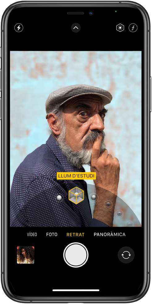 Pantalla de l’app Càmera en el mode Retrat. Al visor, el subjecte es veu nítid i el fons difuminat. El dial per seleccionar els efectes d’il·luminació de Retrat està obert a la part inferior del marc, i hi ha seleccionada l’opció “Llum d’estudi”. A la part superior esquerra de la pantalla hi ha el botó Flaix, al centre de la part superior hi ha botó dels controls de la càmera, i a la part superior dreta els botons per ajustar la intensitat de la il·luminació de Retrat i el control de la profunditat. A la part inferior de la pantalla, d’esquerra a dreta, hi ha el botó visor de fotos i vídeos, el botó per fer una foto i el botó selector de la càmera posterior.