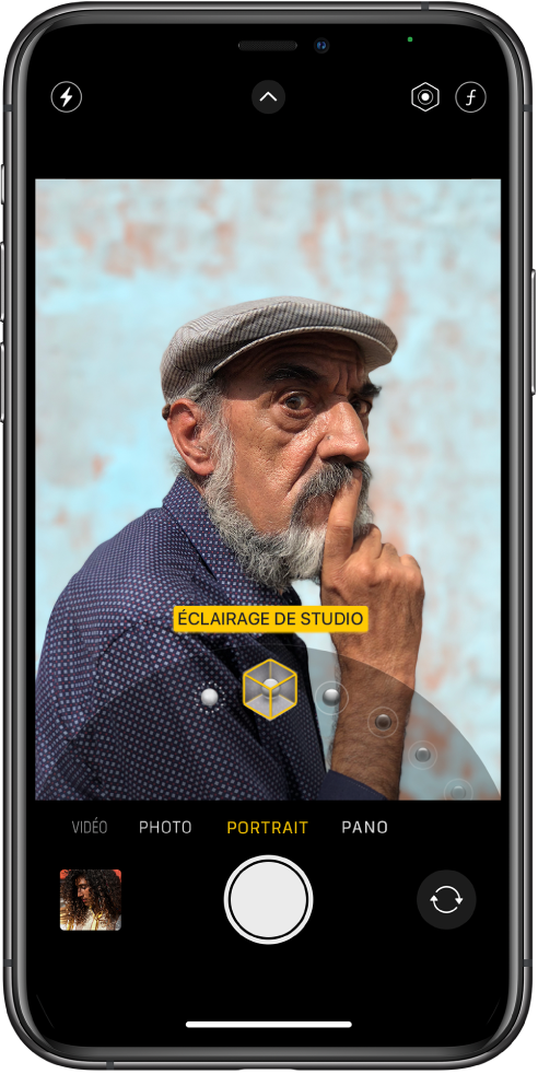 L’écran « Appareil photo » en mode Portrait ; dans le viseur, le sujet est net tandis que l’arrière-plan est flou. Le cadran pour sélectionner les effets d’éclairage Portrait est ouvert en bas de l’image et « Éclairage de studio » est sélectionné. En haut à gauche de l’écran se trouve le bouton Flash, en haut au centre se trouve le bouton « Commandes de l’appareil photo », et en haut à droite de l’écran se trouvent les boutons permettant de régler l’intensité lumineuse du portrait et le contrôle de la profondeur. En bas de l’écran, de gauche à droite, se trouvent le bouton « Visualiseur de photos et vidéos », le bouton « Prendre une photo » et le bouton « Sélecteur de caméra - face arrière ».