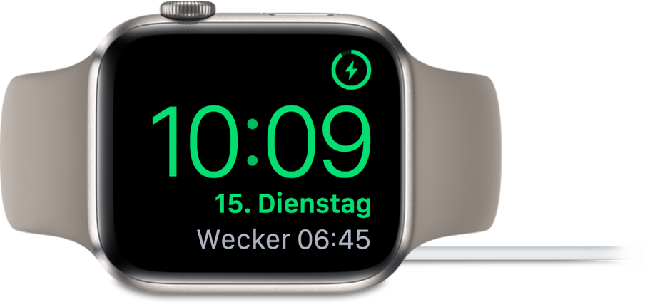 Eine auf die rechte Seite gedrehte Apple Watch, an die das Ladegerät angeschlossen ist und auf deren Display oben rechts das Ladesymbol, darunter die aktuelle Uhrzeit und die Uhrzeit für den nächsten Wecker angezeigt werden.