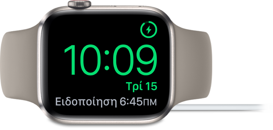 Ένα Apple Watch τοποθετημένο στο πλάι του και συνδεδεμένο στον φορτιστή του. Στην οθόνη εμφανίζεται το σύμβολο φόρτισης στην πάνω δεξιά γωνία, η τρέχουσα ώρα από κάτω η ώρα του επόμενου ξυπνητηριού.