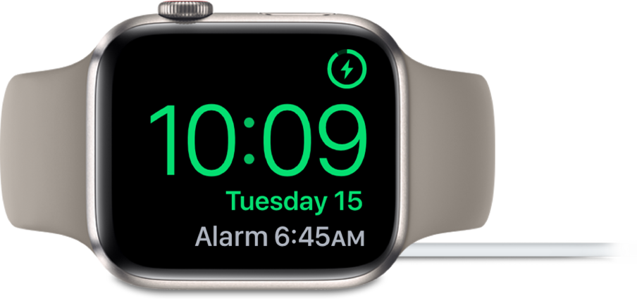 An Apple Watch placed on its side and connected to the charger, with the screen showing the charging symbol in the top-right corner, the current time below that, and the time of the next alarm.