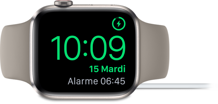Apple Watch placée sur le côté et connectée au chargeur, dont l’écran affiche le symbole de charge en haut à droite, l’heure actuelle en dessous et l’heure de la prochaine alarme.