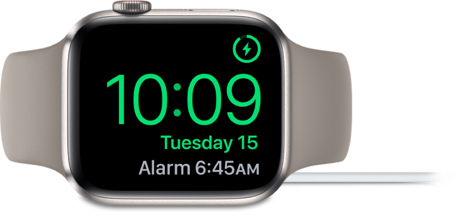 Apple Watch pulkstenis ir novietots uz sāniem un pievienots lādētājam. Tā ekrāna augšējā labajā stūrī ir redzams uzlādes simbols, zem tā ir parādīts pašreizējais laiks, kā arī nākamā modinātāja laiks.