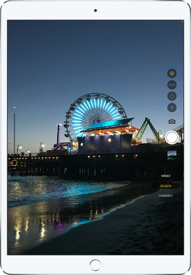 Ilustracja przedstawiająca ekran aplikacji Aparat na iPadzie Pro. Po prawej znajduje się przycisk migawki oraz przyciski przełączania aparatu i wyboru trybu zdjęcia.