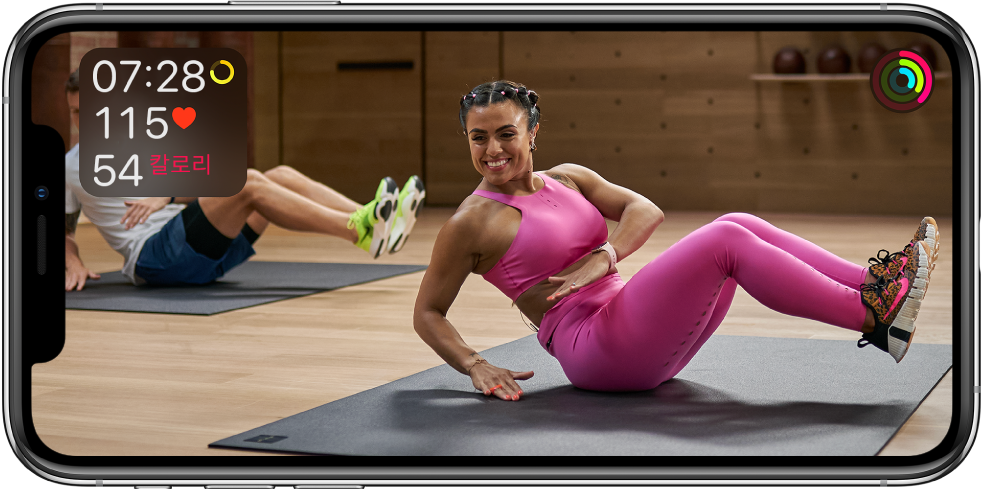 Apple Fitness Plus 운동을 진행하는 트레이너가 나오는 화면. 운동 시간, 심박수 및 소모한 칼로리 정보가 왼쪽 상단에 나타남. 움직이기, 운동하기, 일어서기 목표 진행 상태 고리가 오른쪽 상단에 나타남.