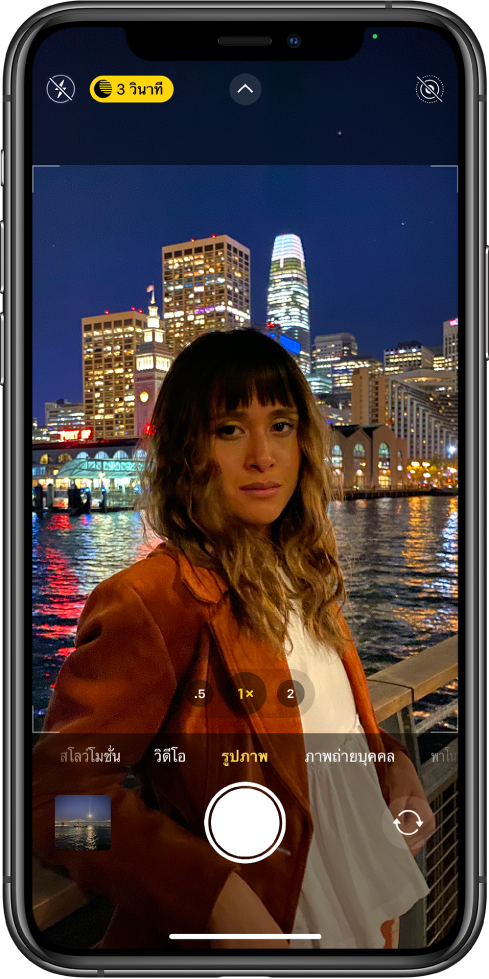 หน้าจอแอพกล้องในโหมดรูปภาพ ปุ่มสำหรับแฟลชและโหมดกลางคืนจะแสดงอยู่ที่มุมซ้ายบนสุดของหน้าจอ แฟลชปิดอยู่และโหมดกลางคืนเปิดใช้อยู่ ปุ่มตัวควบคุมกล้องอยู่ตรงกึ่งกลางด้านบนสุด และปุ่ม Live Photos อยู่ตรงมุมขวาบนสุด ด้านล่างสุดของหน้าจอจากซ้ายไปขวา คือ ปุ่มหน้าต่างแสดงรูปภาพและวิดีโอ ปุ่มถ่ายรูป และปุ่มตัวเลือกกล้องด้านหลังและด้านหน้า