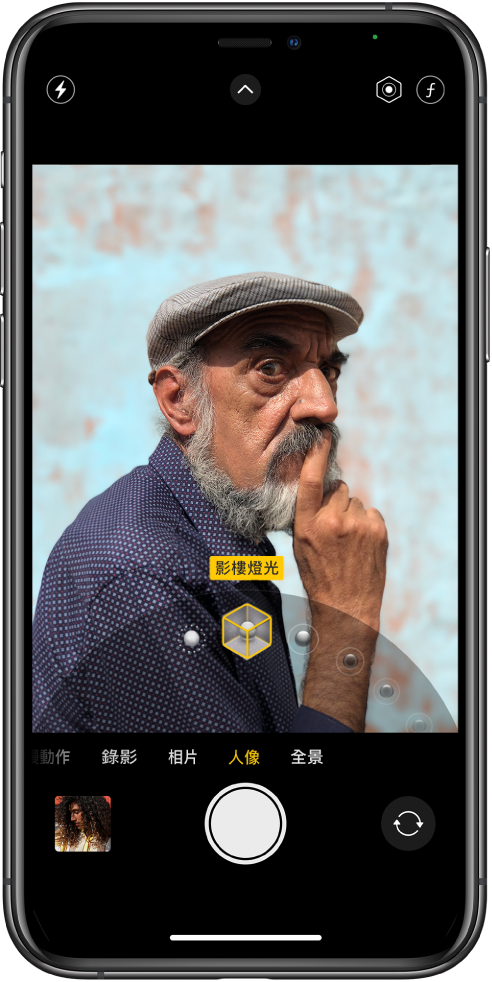 「相機」畫面中的「人像」模式；在觀景器中，主體清晰而背景模糊。用於選擇「人像燈光」效果的刻度盤已在影格下方開啟，並已選擇「影樓燈光」。螢幕左上方是「閃光燈」按鈕，中間上方是「相機控制項目」，而右上方是調整「人像」燈光強度及景深控制的按鈕。螢幕底部由左至右為：「相片和影片檢視器」按鈕、「影相」按鈕和「相機選擇器後置鏡頭」按鈕。