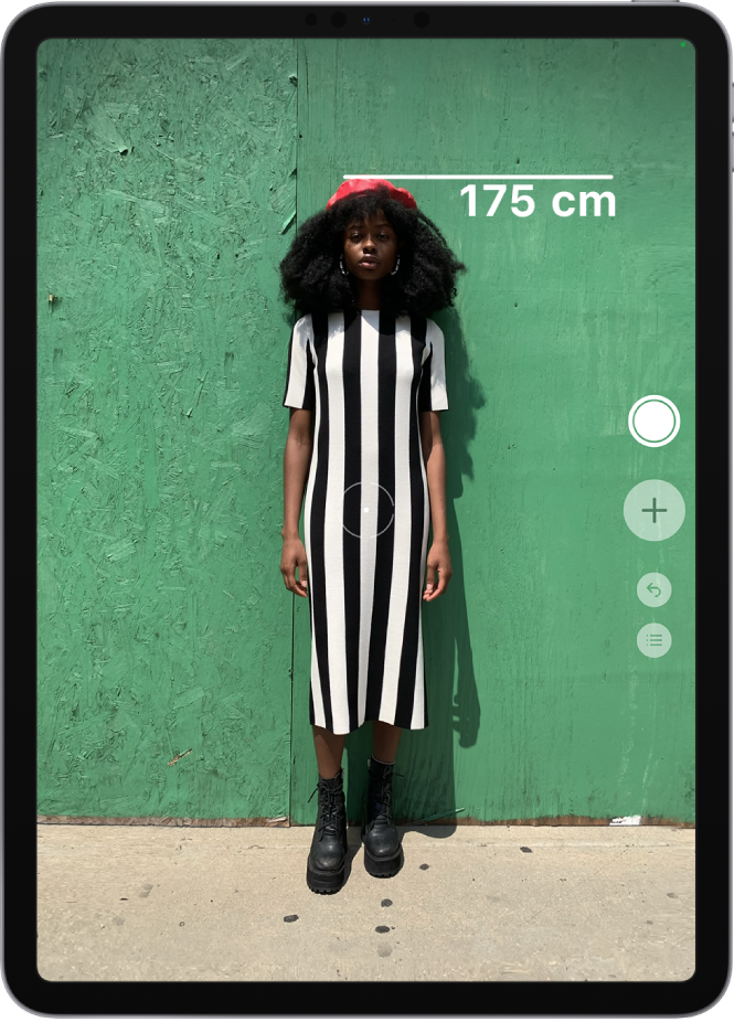 Une personne en train d’être mesurée, la taille s’affichant au-dessus de sa tête. Le bouton « Prendre une photo » est actif sur le bord droit, afin de prendre une photo de la mesure. L’indicateur vert « Caméra en cours d’utilisation » apparaît en haut à droite.