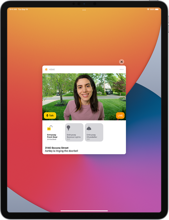 На экране iPad отображается уведомление из приложения «Дом». На нем изображен человек у входной двери и кнопка «Говорите» слева. Ниже расположены кнопки аксессуаров для управления входной дверью и освещением при входе. Внизу указан адрес дома вместе со словами «Саша звонит в дверь». Кнопка «Закрыть» находится в правом верхнем углу уведомления.