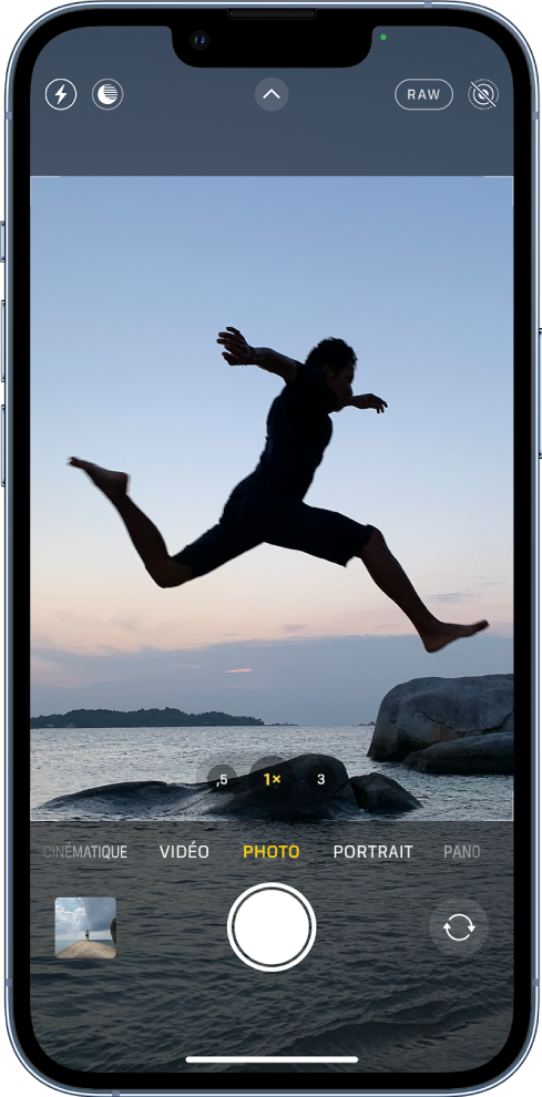 L’écran Appareil photo en mode Photo, avec les autres modes à gauche et à droite sous le viseur. Les boutons pour le flash, le mode Nuit, les commandes de l’appareil photo, le format ProRaw et le mode Live Photo apparaissent en haut de l’écran. En dessous des différents modes d’appareil photo, de gauche à droite, se trouvent le bouton « Visualiseur de photos et vidéos », le bouton « Prendre une photo » et le bouton « Sélecteur de caméra - face arrière ».