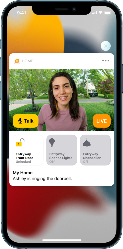 A notification from Home is on the iPhone screen. It shows the picture of a person at the front door with a Talk button at the left. Below are accessory buttons for the front door and entryway lights. The words “Ashley is ringing the doorbell.” A Close button is at the top right of the notification.