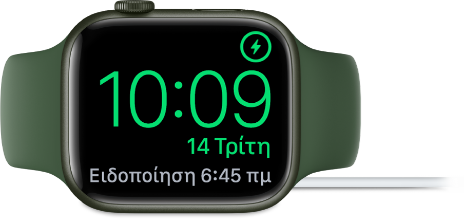 Ένα Apple Watch τοποθετημένο στο πλάι του και συνδεδεμένο στον φορτιστή του. Στην οθόνη εμφανίζεται το σύμβολο φόρτισης στην πάνω δεξιά γωνία, η τρέχουσα ώρα από κάτω η ώρα του επόμενου ξυπνητηριού.