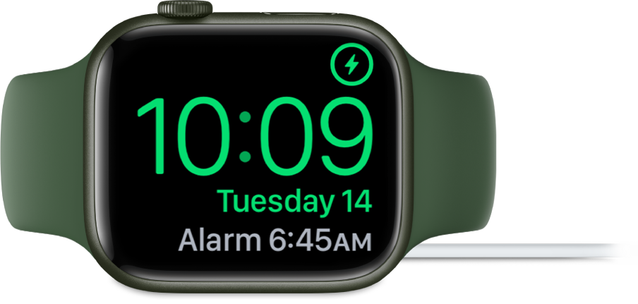 An Apple Watch placed on its side and connected to the charger, with the screen showing the charging symbol in the top-right corner, the current time below that, and the time of the next alarm.