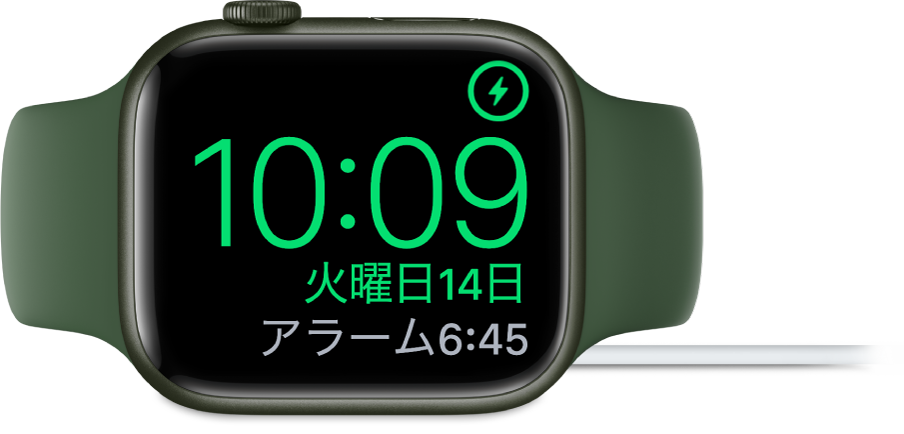 横向きに置かれ、充電器に接続されているApple Watch。画面には、右上隅に充電中のマーク、その下に現在時刻と、次のアラーム時刻が表示されています。