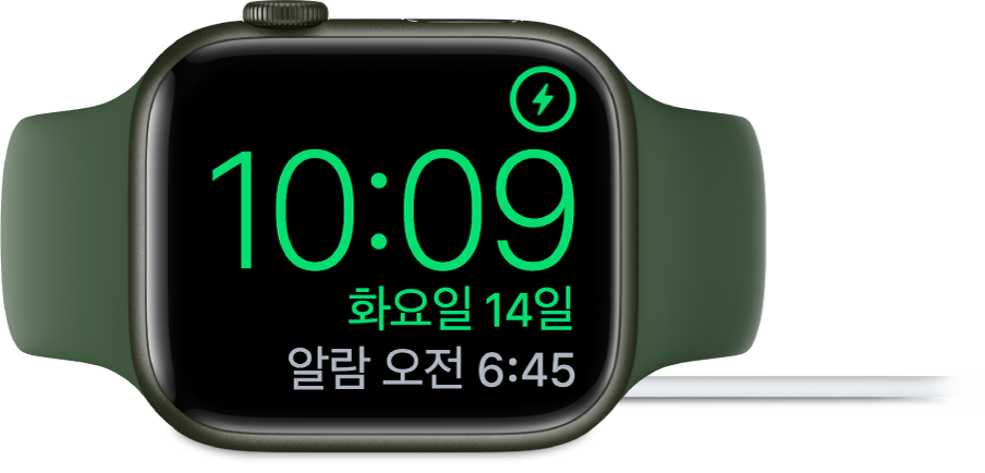 Apple Watch가 옆으로 누워있고 충전기에 연결되어 있음. 화면의 오른쪽 상단 모서리에는 충전 기호가 있고 그 아래에는 현재 시간과 다음 알람 시간이 있음.