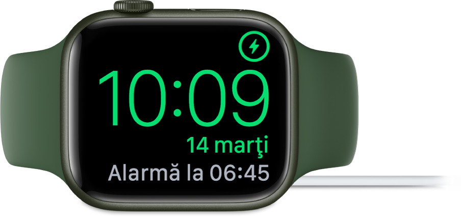 Un Apple Watch așezat pe muchie și conectat la încărcător, pe ecran fiind afișat simbolul de încărcare în colțul din dreapta sus, ora actuală dedesubt și ora alarmei următoare.